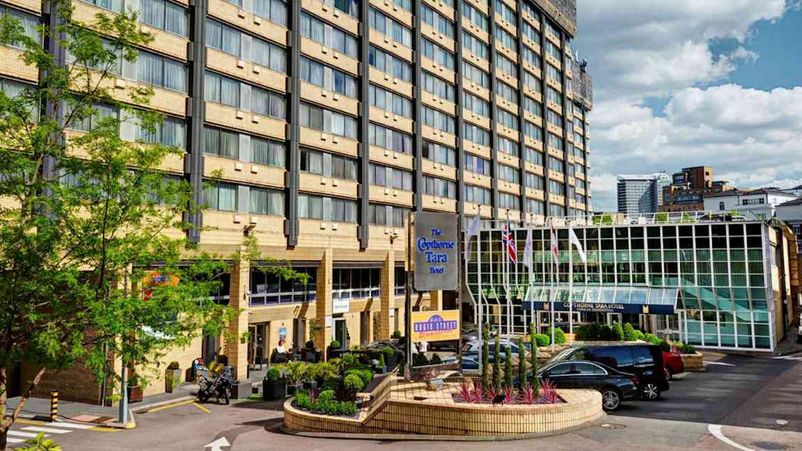 Hotellbyggnad med flera våningar, en glasentré och en parkeringsplats framför. (AI-genererad text)