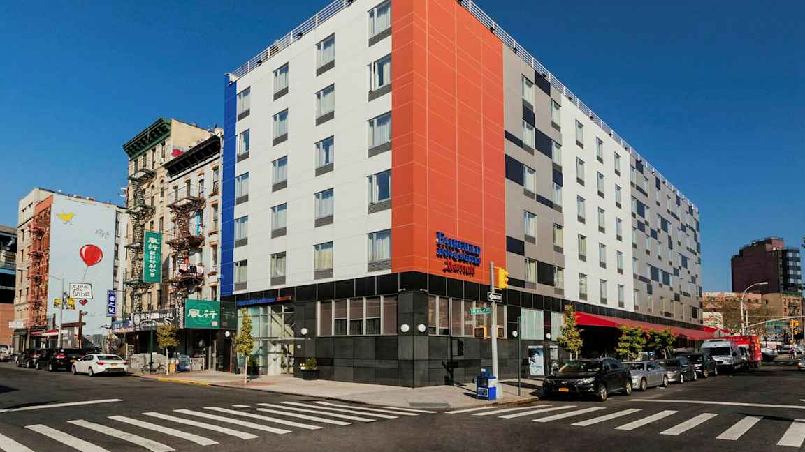En modern byggnad i ett hörn av ett stadsområde, med en livlig gata och trafik samt omgivande äldre byggnader. (AI-genererad text)