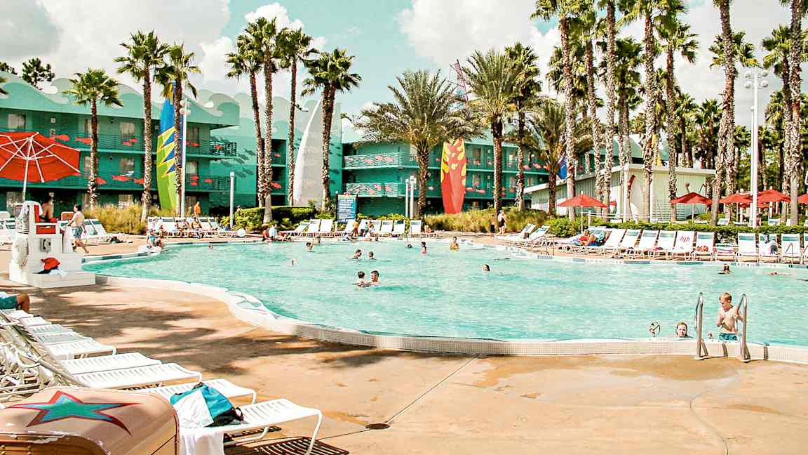 En utomhuspool omgiven av solstolar, parasoller och palmer. I bakgrunden syns en byggnad med färggranna detaljer. (AI-genererad text)