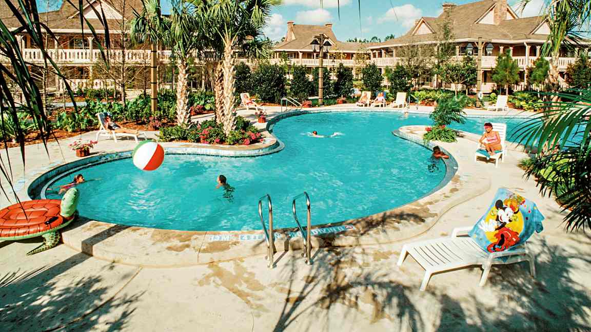 En utomhuspool omgiven av palmer och solstolar, med personer som badar och leker i vattnet. Byggnader syns i bakgrunden. (AI-genererad text)