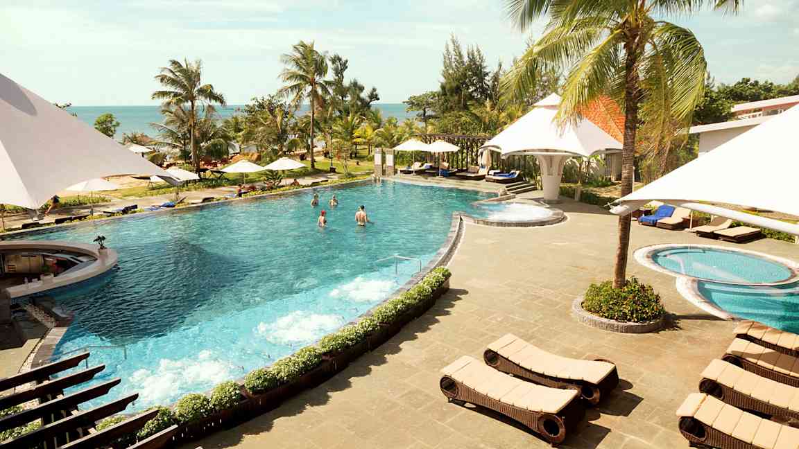 En utomhuspool med omgivande solstolar och paraplyer vid ett tropiskt semesterområde. Palmträd och hav i bakgrunden. (AI-genererad text)