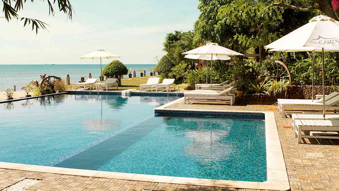 En utomhuspool omgiven av solstolar, parasoller och grönska nära stranden. (AI-genererad text)