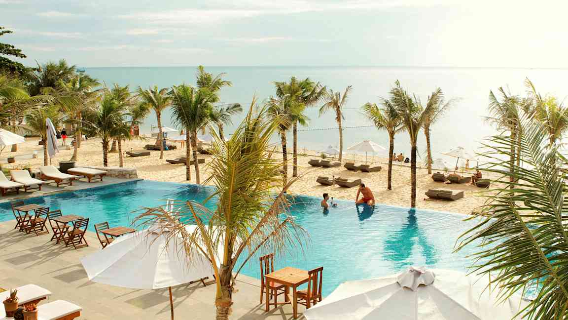 En pool med solstolar och utemöbler i förgrunden, palmer, sandstrand och hav i bakgrunden. (AI-genererad text)