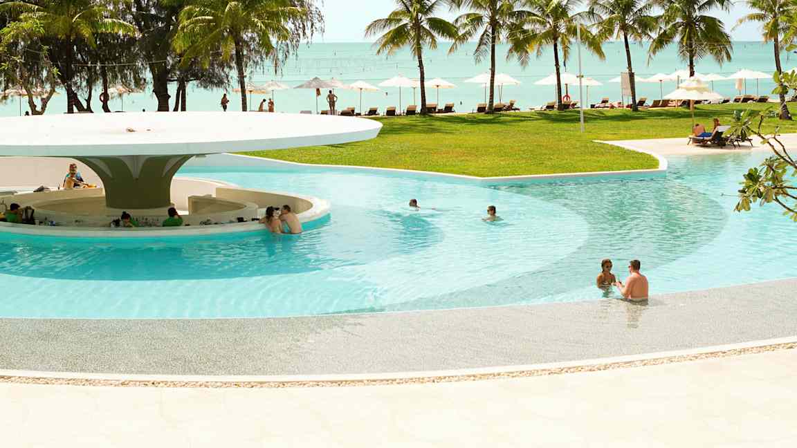 En pool med sittplatser i vattnet och en bar under ett stort tak. Bakom poolen finns gräsmatta, solstolar, parasoller, palmer och utsikt över havet. (AI-genererad text)