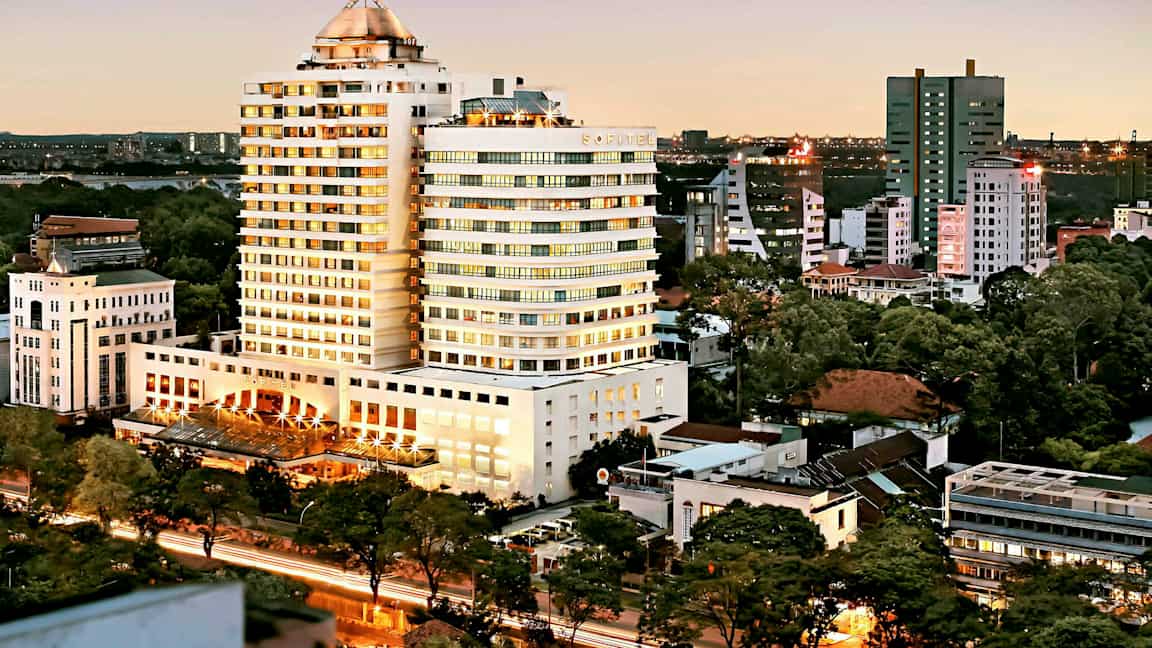Vy över en stad under kvällstid med en stor byggnad i centrum, omgiven av mindre byggnader och träd. (AI-genererad text)