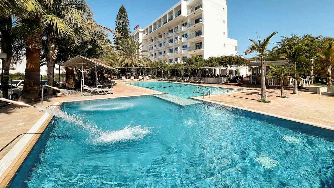 En hotellpool omgiven av palmer och ett vitt byggnad i bakgrunden. (AI-genererad text)