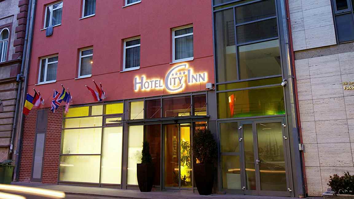 Utanför en hotellbyggnad med en skylt som lyder "Hotel City Inn" samt flera flaggor vid entrén. (AI-genererad text)