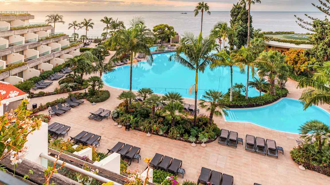 Bild på ett hotellområde med en stor swimmingpool omgiven av solstolar och tropiska växter. I bakgrunden syns havet och en solnedgång. (AI-genererad text)