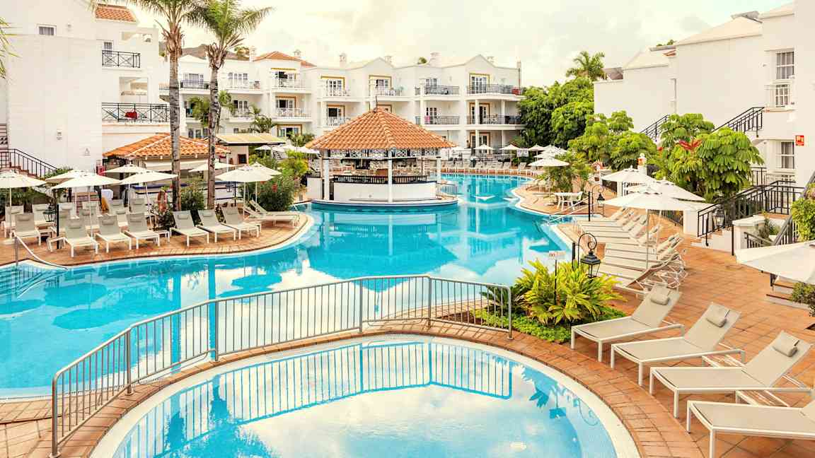 En pool med omgivande solstolar och parasoller samt vita byggnader i bakgrunden. (AI-genererad text)