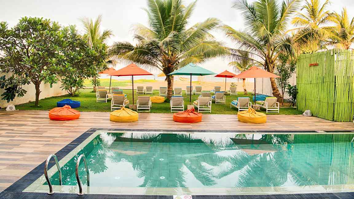 En pool med solsängar och färgglada parasoller i en trädgård omgiven av palmer. (AI-genererad text)