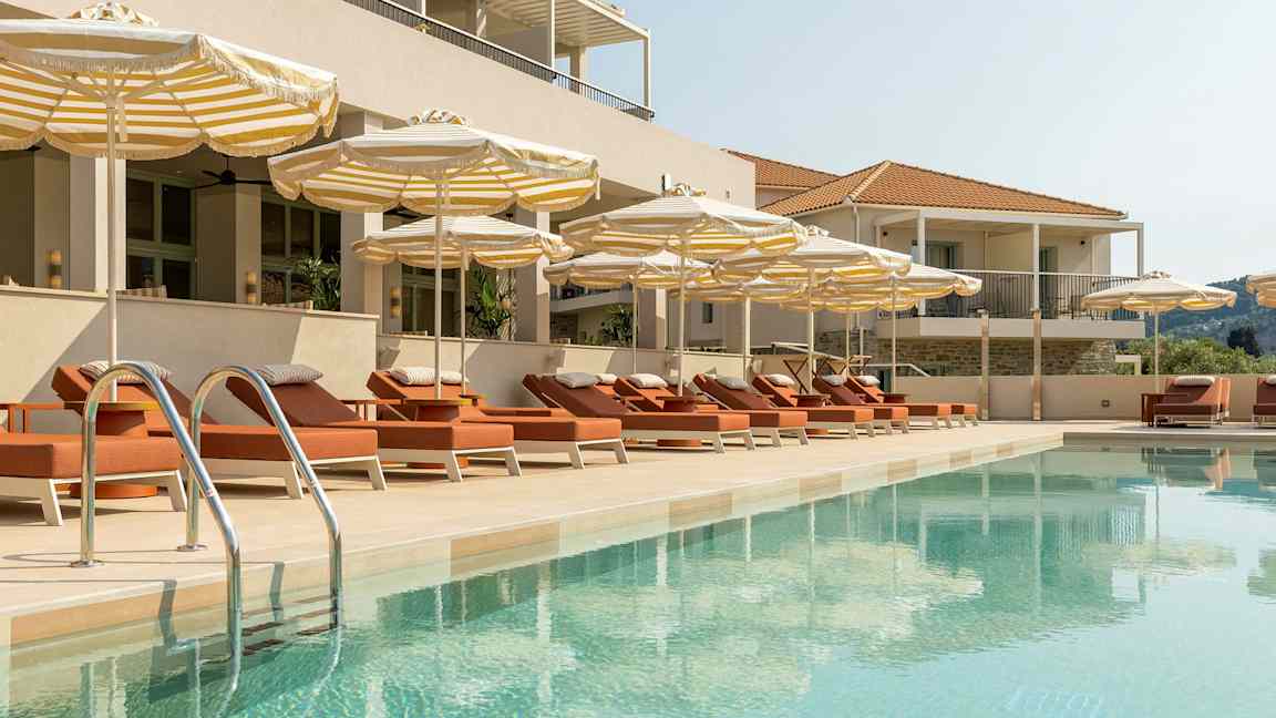 En pool omgiven av solstolar och parasoller vid en byggnad. (AI-genererad text)