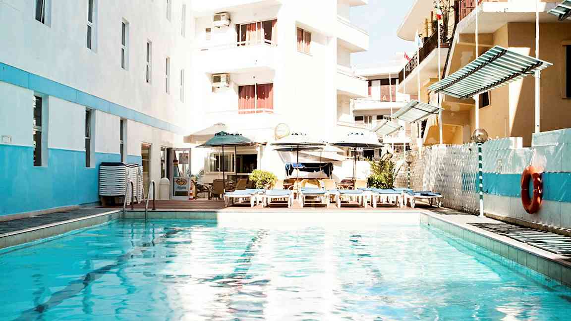 En utomhuspool omgiven av hotellbyggnader och solstolar. (AI-genererad text)