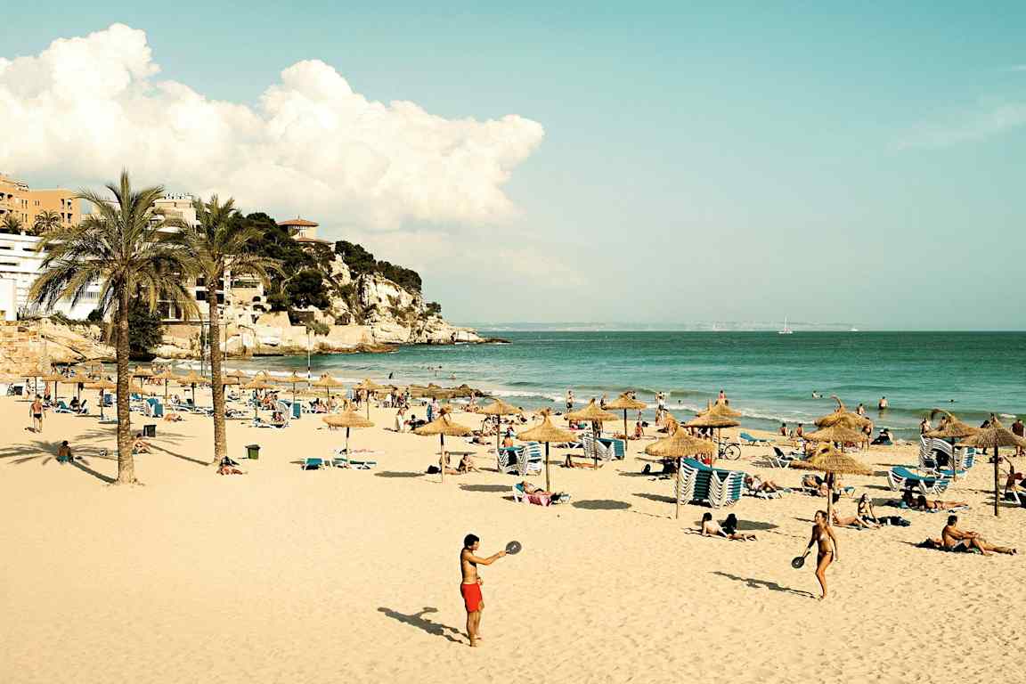 En sandstrand med parasoller, solstolar och människor som njuter av solen och havet. Längre bort syns en klippa med byggnader. (AI-genererad text)