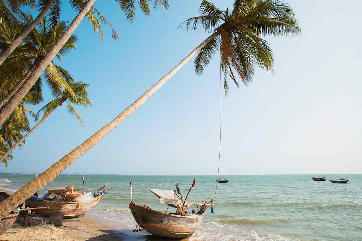 Utsikt över en tropisk strand med palmer och traditionella båtar vid vattnet. (AI-genererad text)