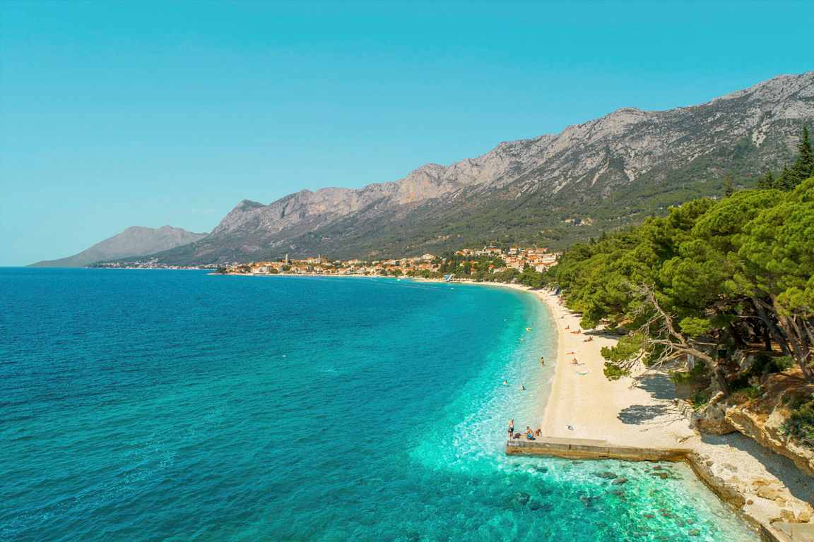 Vy över en klapperstensstrand med turkost vatten och grönskande träd. Berg och en by syns i bakgrunden. (AI-genererad text)