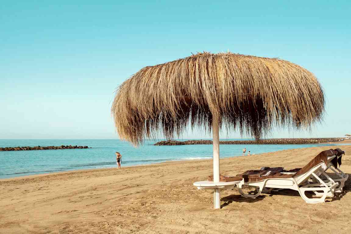 Bild av en sandstrand med två solstolar under ett parasoll av halm samt människor som går längs vattnet. (AI-genererad text)