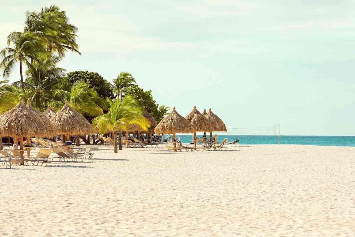 En strand med parasoll gjort av palmblad, solstolar och ett nät för strandvolleyboll. (AI-genererad text)
