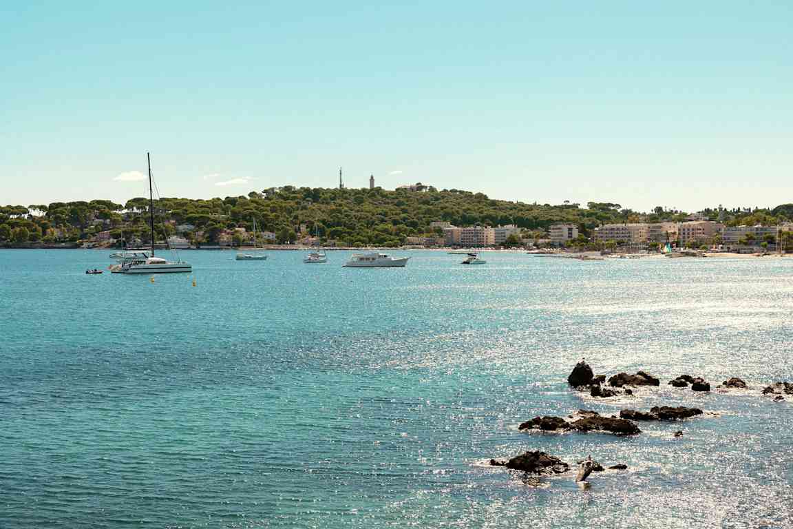 Vy över en kustlinje med klarblått vatten, flera båtar och segelbåtar i närheten av en grönskande strand och en stad. (AI-genererad text)