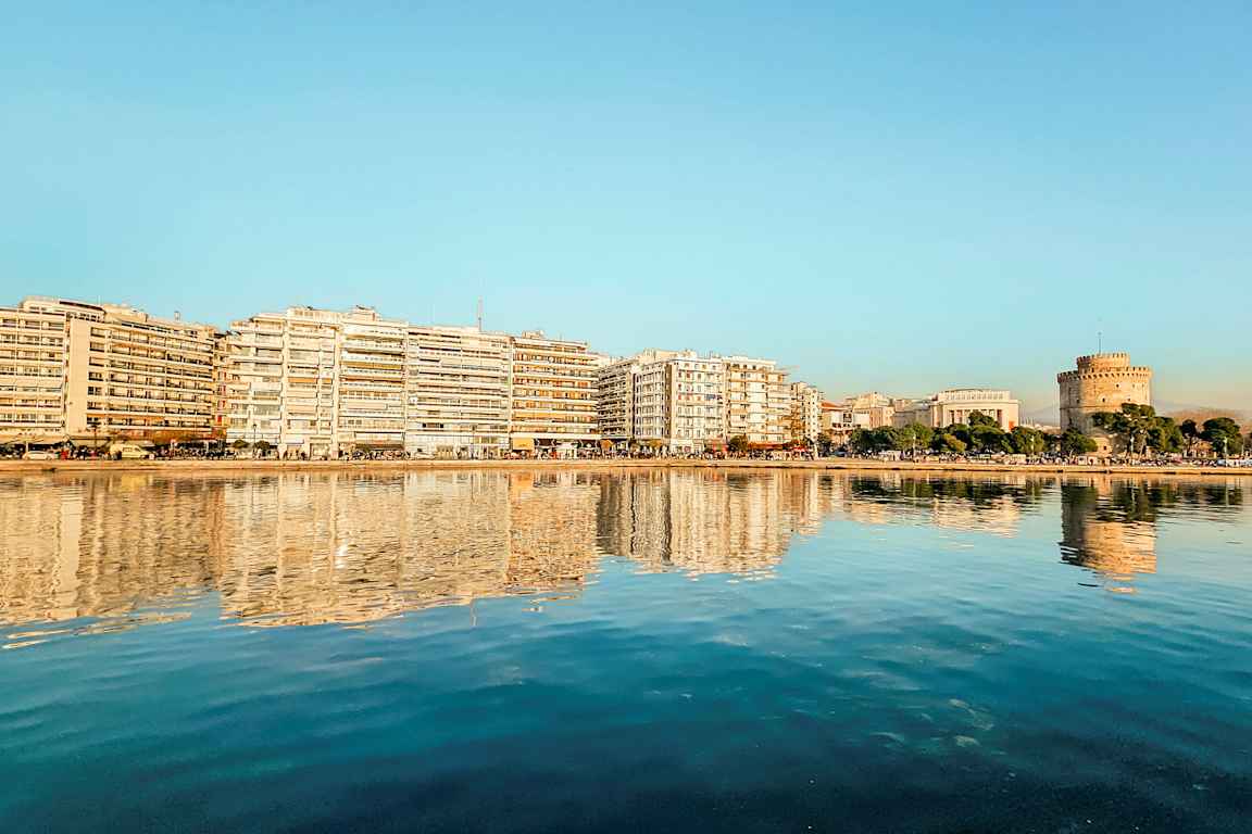 En stad vid vattnet med byggnader och ett runt torn reflekterade i det lugna vattnet. (AI-genererad text)