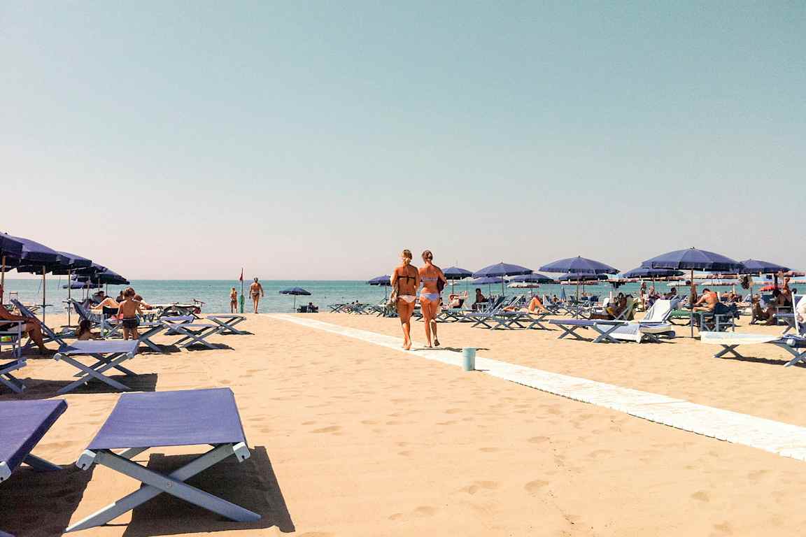 En sandstrand med blå solstolar och parasoll. Människor solar och går på stranden med en klar blå himmel i bakgrunden. (AI-genererad text)
