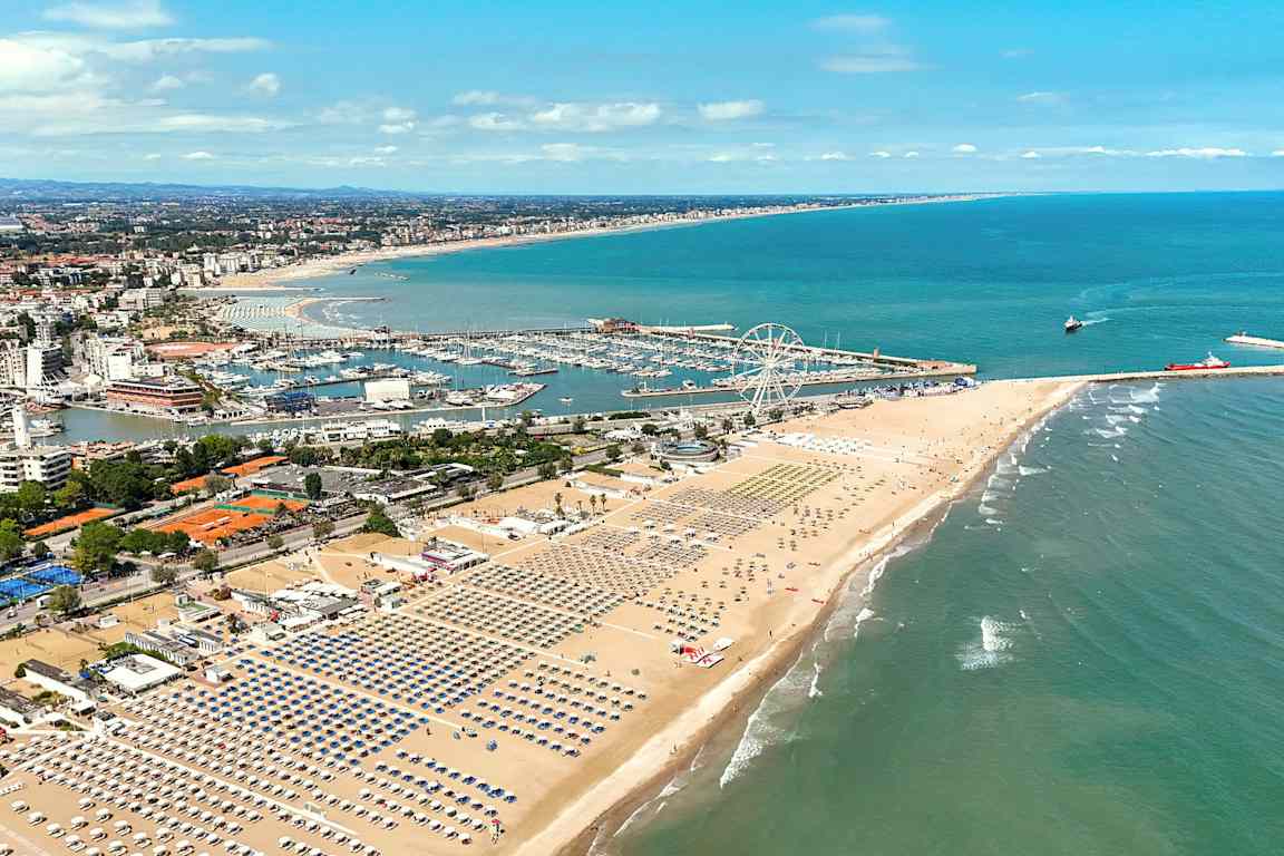 En vy över en bred sandstrand med rad efter rad av solstolar och parasoller. I bakgrunden syns en småbåtshamn och en nöjesfältsliknande miljö med ett stort pariserhjul. Havet breder ut sig till höger. (AI-genererad text)