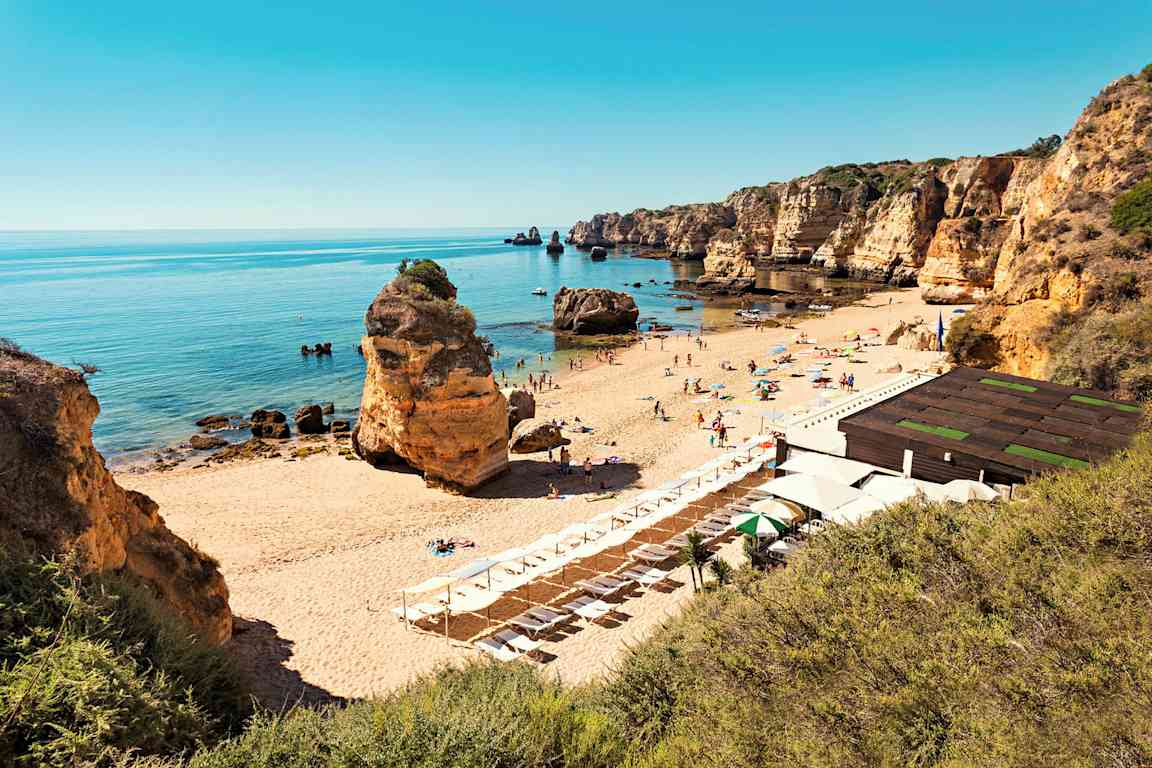 En sandstrand med solstolar och parasoller, omgiven av klippformationer och ett stilla hav. (AI-genererad text)