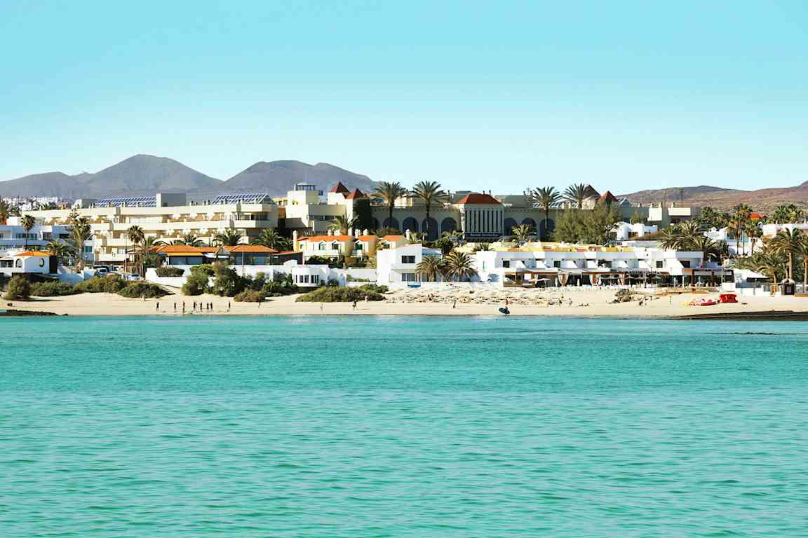 En strand med vit sand och turkost vatten framför en rad av byggnader och palmer. (AI-genererad text)