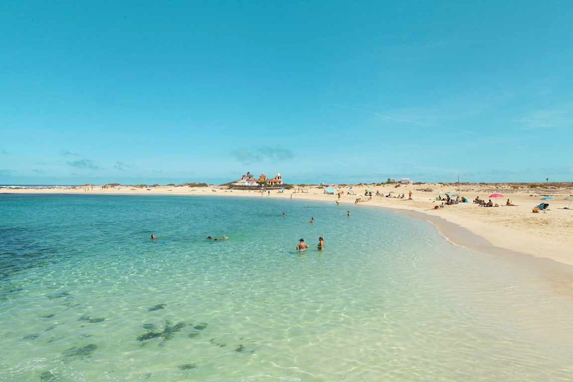 En strand med solbadande människor, några simmar i det kristallklara turkosa vattnet. Horisonten visar ett öde landskap med några byggnader. (AI-genererad text)