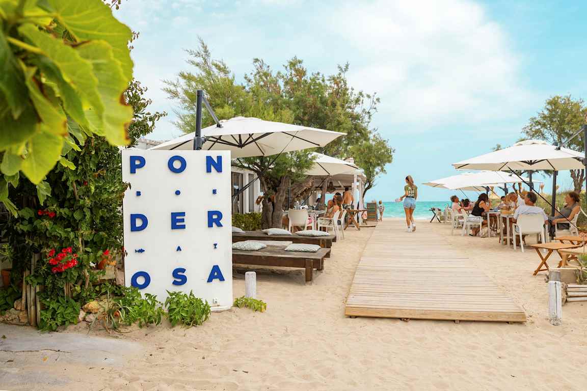 En restaurang vid stranden med bord och parasoller, en trägång leder mot vattnet. (AI-genererad text)