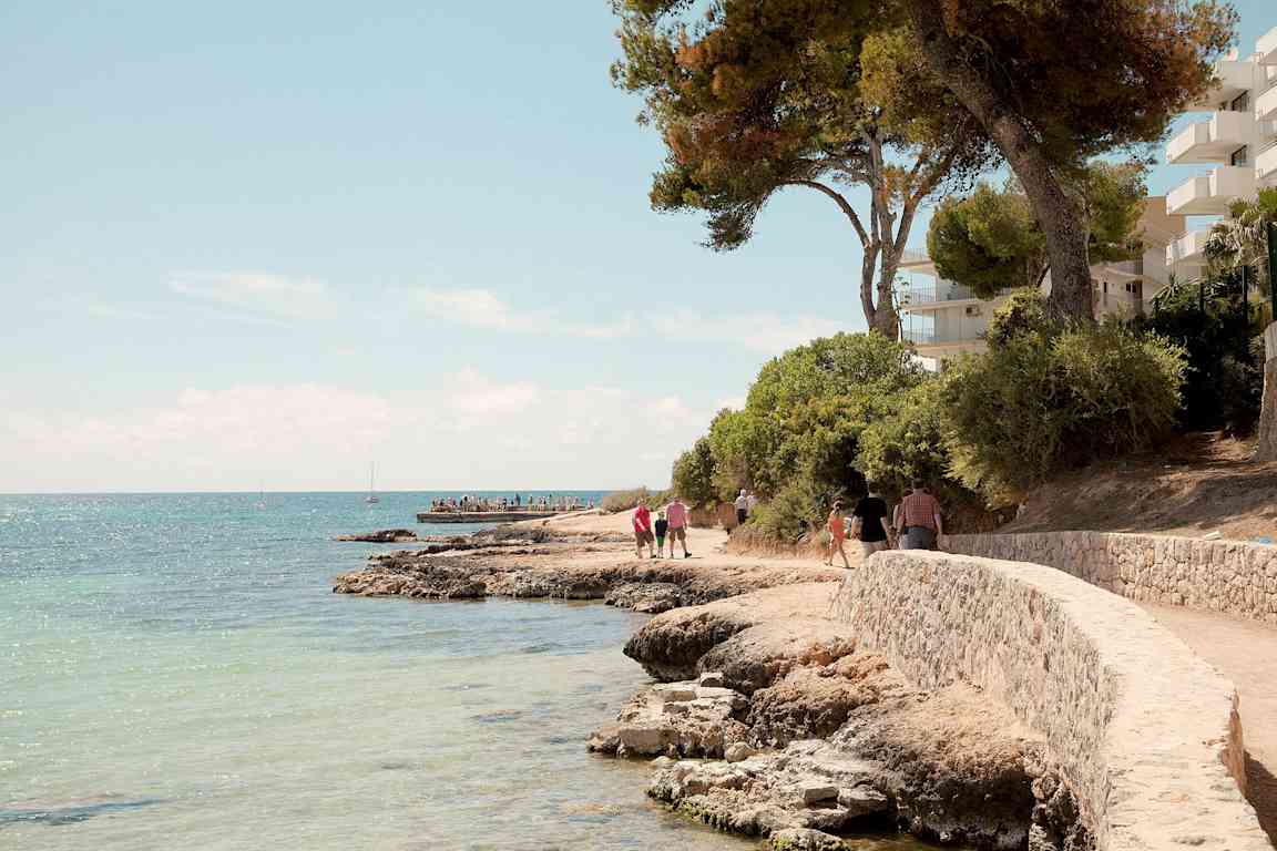Stenstrand med slingrande gångväg intill havet, omgivet av träd och byggnader. (AI-genererad text)
