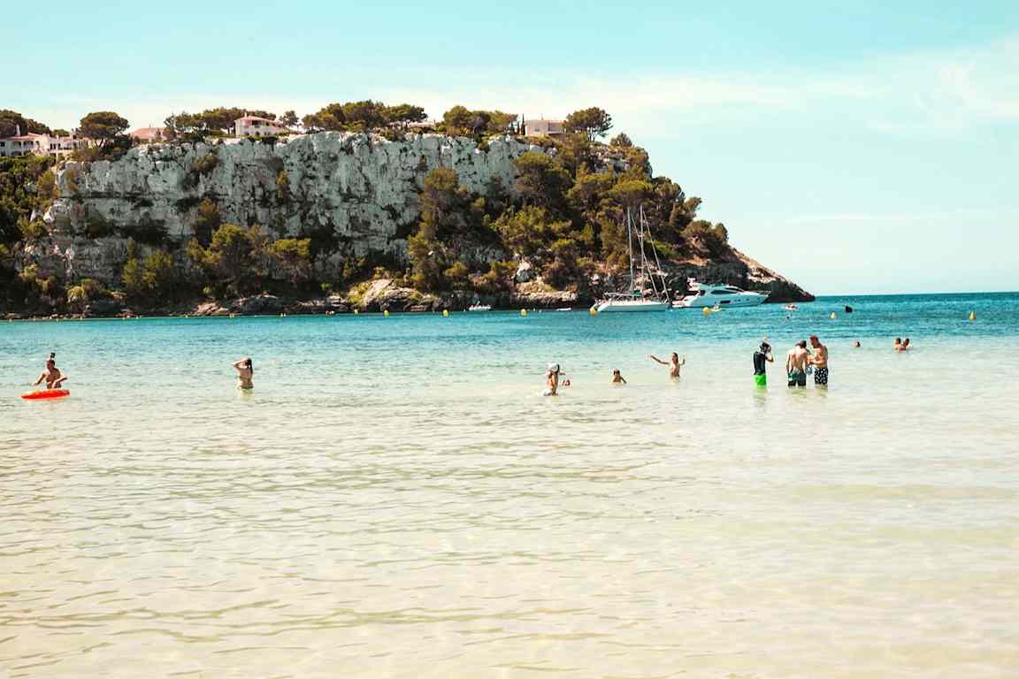 En strand med människor i vattnet framför en kulle med klippor och några hus samt båtar i bakgrunden. (AI-genererad text)