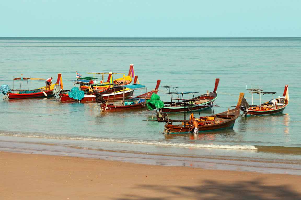 Flera färgglada båtar ligger förtöjda på vatten nära en sandstrand. (AI-genererad text)