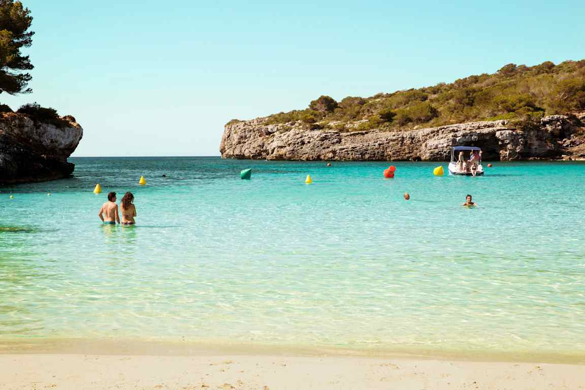 En sandstrand med människor i det lugna, turkosa vattnet. I bakgrunden syns en båt och klippor täckta av grönska. (AI-genererad text)