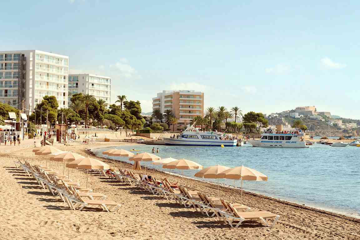 Strand med solstolar och parasoller, hotellbyggnader i bakgrunden och båtar på vattnet. (AI-genererad text)