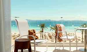Utsikt från en balkong över en strand med solstolar och parasoller, palmer samt hav och berg i fjärran. (AI-genererad text)