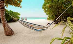 Person som vilar i en hängmatta på en sandstrand med träd och hav i bakgrunden. (AI-genererad text)