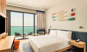 Ett hotellrum med en vitbäddad dubbelsäng, en TV på en bänk, och en sittgrupp intill en balkong med utsikt över havet. (AI-genererad text)