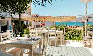 En uteservering med vita trästolar och bord, dekorerad med blommor, och en utsikt över havet. (AI-genererad text)