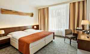 Ett hotellrum med en dubbelsäng som har ett orange överkast och två vita kuddar. Det finns nattduksbord med lampor på båda sidor och ett stort fönster med gardiner i bakgrunden. Ett skrivbord med en stol och diverse föremål står till höger. (AI-genererad text)