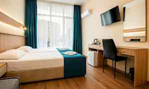 Ett hotellrum med en dubbelsäng, skrivbord, stol, väggmonterad TV och stora fönster med mörkblå gardiner. (AI-genererad text)