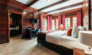 Hotellrum med träinredning, en bred säng med vita och färgglada kuddar, röda gardiner och fönster med utsikt. (AI-genererad text)