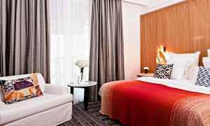 Hotellrum med säng, kudde med mönster, röd filt, stol och bord med blommor. (AI-genererad text)