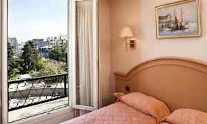 Ett hotellrum med en säng och en öppen balkongdörr som visar utsikt över en stad och grönområde. (AI-genererad text)