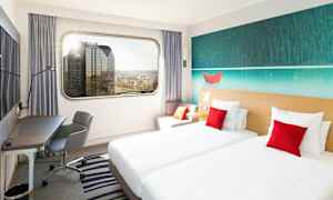 Ett hotellrum med två enkelsängar, en arbetshörna och en stor fönstervy över stadens skyline. (AI-genererad text)