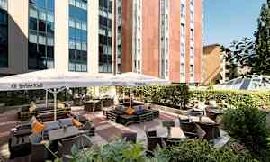 Bild på en uteservering med bord, stolar och parasoll vid en byggnad. (AI-genererad text)