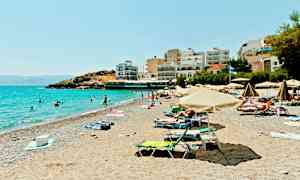 En solig strand med parasoller, människor som badar och byggnader vid strandkanten. (AI-genererad text)