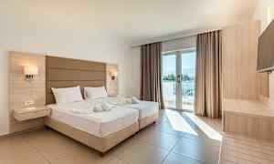 Ett ljust hotellrum med två sängar, en balkongdörr med utsikt och möbler av trä. (AI-genererad text)