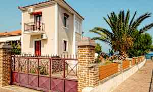 En tvåvåningsbyggnad med balkong omgiven av en stenmur och grind, bredvid en palm och blommande buskar. (AI-genererad text)