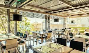 En restaurang med flera dukade bord och stolar, med fönster mot en stadsvy. (AI-genererad text)