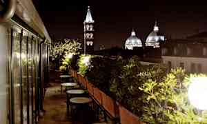 En uteplats på taket med bord och stolar, omgiven av gröna växter, med kyrkkupoler och ett klocktorn som syns i bakgrunden på kvällen. (AI-genererad text)